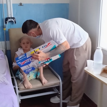 Un hombre con mascarilla sostiene una caja de juguetes frente a un niño sentado en un sillón junto a un suero