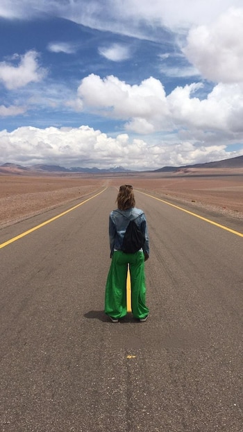 Una persona de espaldas con chaqueta vaquera y pantalón verde está en medio de una carretera asfaltada en un paisaje desértico