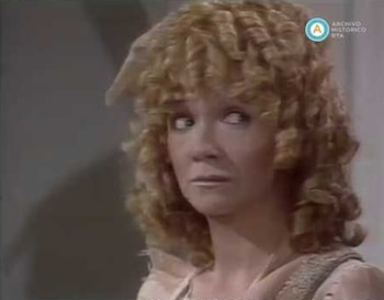 Primer plano de una mujer con cabello rubio rizado que mira hacia la derecha. La mujer tiene los labios maquillados. En la esquina superior derecha, se observa un logo