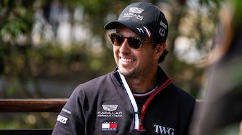 Primer plano de un hombre sonriente con gafas de sol, gorra negra con el logo de Cadillac Formula 1 Team y una chaqueta oscura con parches de patrocinadores.