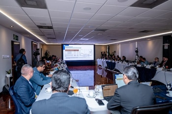 Cinco hombres con traje están sentados en una mesa larga con monitores, micrófonos y carteles de identificación