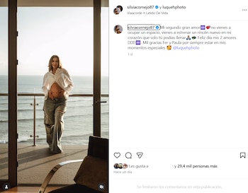 Mujer embarazada de pie en un balcón con vista al mar, vestida con camisa blanca abierta y pantalones vaqueros rotos, manos sobre el vientre