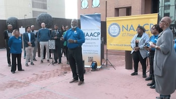 Un hombre habla ante un grupo de personas frente a dos carteles de la organización NAACP en una plaza al aire libre