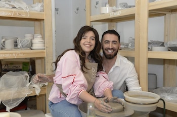 Una mujer y un hombre sonríen frente a un torno con arcilla, rodeados por estantes de madera con recipientes de cerámica
