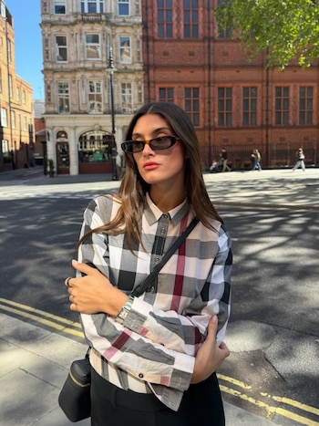 Mujer joven con cabello castaño y gafas de sol, vistiendo camisa a cuadros y pantalones oscuros, de pie en una calle de Londres con edificios antiguos de ladrillo