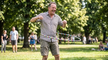 Un hombre con camisa estampada y pantalón corto gira un hula hula multicolor en su cintura en un parque con árboles y personas.