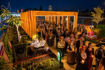 Personas con maquillaje de calavera y ropa tradicional caminan de noche por una estructura de madera con velas encendidas.