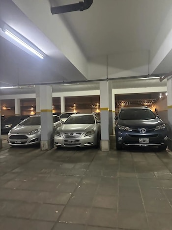 Estacionamiento interior iluminado con luces fluorescentes, varios coches aparcados entre columnas de concreto grises y una tubería negra en el techo