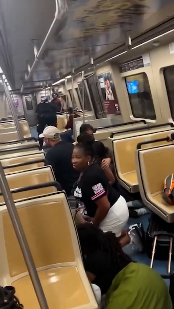 Interior de un vagón de tren con pasajeros agachados o en el suelo. Los asientos son beige y hay barras de sujeción plateadas. Se ven varias personas en el pasillo