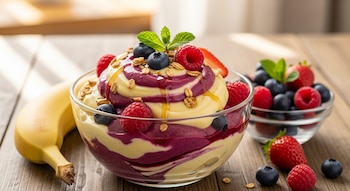 Un bol de helado de plátano y frutos rojos con granola y hojas de menta, rodeado de plátanos, arándanos, fresas y frambuesas, en una mesa de madera.