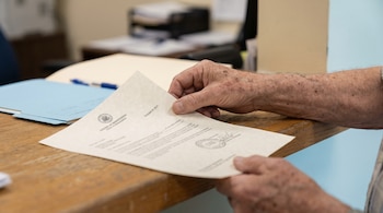Mano de persona mayor sosteniendo un certificado oficial con sellos. Mostrador de madera clara, carpeta azul y bolígrafo al fondo desenfocado.