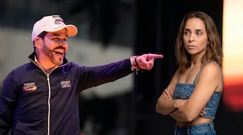 Un hombre con gorra y chaqueta oscura sonríe y señala a una mujer con top azul de mezclilla y brazos cruzados, con expresión seria.