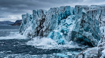 Pared de hielo agrietada cae al mar entre olas y espuma, con un cartel abajo a la derecha que lleva el texto ¡EMERGENCIA CLIMÁTICA!.