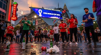 Multitud de aficionados bebiendo en una calle mojada al atardecer frente al Mercedes-Benz Stadium de Atlanta con un cartel de la Copa Mundial FIFA 2026. Hay basura en el suelo.