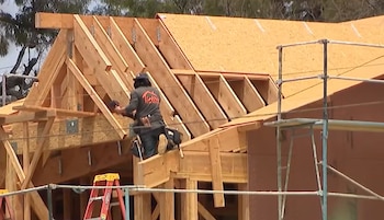 Hombre sobre tejado de madera en construcción con herramienta, rodeado de andamios y una escalera roja bajo un cielo claro