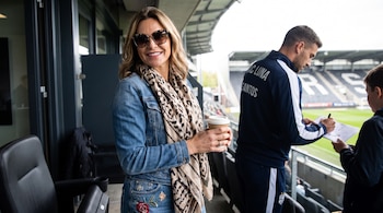 Una mujer con gafas de sol, chaqueta vaquera y pañuelo sostiene un vaso de café; un hombre con chándal firma un autógrafo a un niño en un estadio.