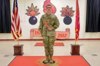 Un hombre en uniforme militar camuflado sostiene un tubo rojo entre la bandera de Estados Unidos y un estandarte rojo en una sala