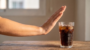 Antebrazo y mano con palma abierta haciendo un gesto de detención frente a un vaso con refresco, hielo y burbujas sobre una mesa de madera.
