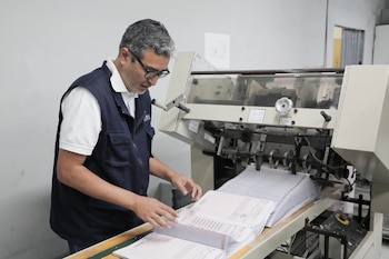Un hombre canoso con gafas y un chaleco azul oscuro manipula pilas de papel blanco en una máquina industrial de color beige en un entorno de taller
