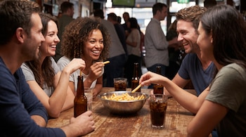 Cinco personas, tres mujeres y dos hombres, sentadas en una mesa de madera. En el centro hay un tazón de papas fritas y varias bebidas en botellas y vasos.