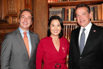 Tres personas posando, dos hombres y una mujer. Un hombre de traje gris, una mujer de vestido rojo, y otro hombre de traje oscuro con pin de bandera. Fondo con libros