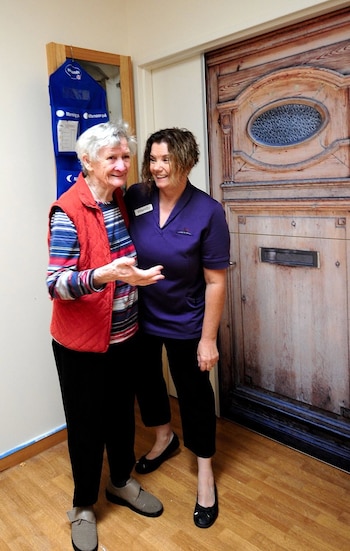 Dos mujeres de pie, una mayor con chaleco rojo y camisa de rayas, otra con uniforme morado y placa que indica "Wendy". Detrás, una puerta de madera tallada y un organizador azul en la pared