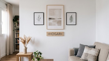 Pared blanca con tres cuadros, cartel de madera con el texto «HOGAR», un sofá con cojines, mesas auxiliares y plantas decorativas.
