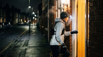 Persona con gorro, linterna frontal, chaqueta reflectante y mochila abriendo la puerta de una casa por la noche. Se ve vapor de aliento y una calle iluminada al fondo.