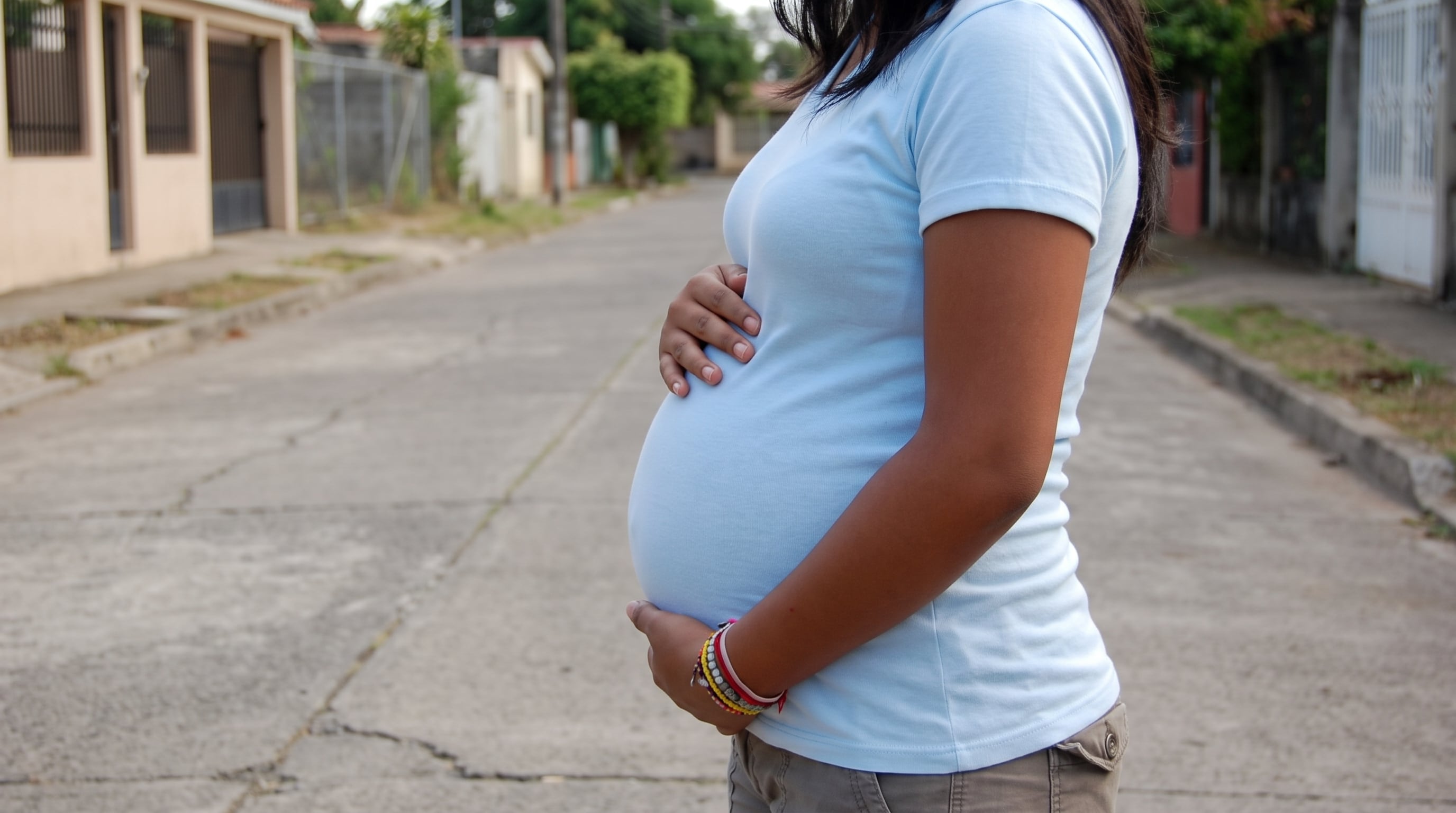 Una mujer embarazada con camiseta azul clara muestra su abdomen de perfil en una calle residencial. (Imagen Ilustrativa Infobae)