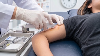 Manos con guantes aplican un dispositivo en el brazo de una persona recostada, junto a una bandeja con instrumental médico.