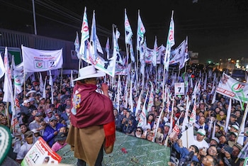 Vista trasera de un orador con sombrero y poncho, dirigiéndose a una gran multitud que sostiene banderas de 'Juntos por el Perú' en un mitin nocturno