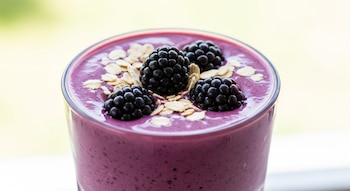 Un licuado cremoso violeta intenso en vaso alto, adornado con cuatro moras frescas y copos de avena, sobre un fondo desenfocado claro.