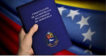 Primer plano de una mano que sostiene la Constitución de la República Bolivariana de Venezuela, con el escudo nacional, sobre una bandera venezolana