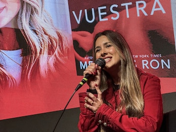Mercedes Ron, mujer de cabello largo y rubio oscuro, sonríe y sostiene un micrófono con las dos manos frente a una pantalla roja