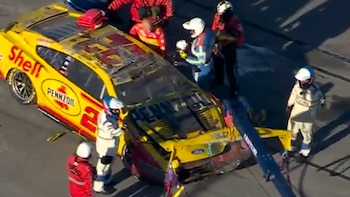 Un automóvil de carreras amarillo y rojo dañado sobre la pista rodeado por operarios de asistencia con cascos y una grúa de remolque