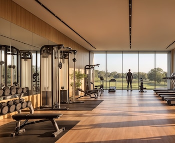 Interior de un gimnasio con máquinas, mancuernas y una persona parada frente a un gran ventanal con vista a un parque