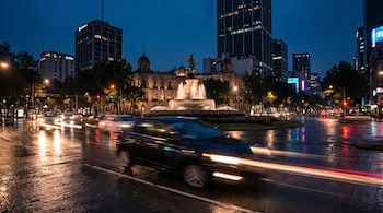 Coche oscuro con estelas de luz en una avenida urbana mojada de noche. Al fondo, la estatua de la Diana Cazadora y edificios altos iluminados por la ciudad.