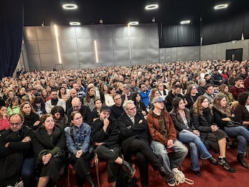 Multitud de personas sentadas en un amplio auditorio con paredes grises y luces circulares en el techo oscuro, atentas al evento