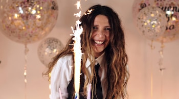 Mujer de cabello castaño y largo sonriendo, con una bengala encendida al frente y globos de fiesta con confeti en el fondo desenfocado.