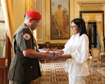 Hombre en uniforme militar verde y boina roja entrega pila de documentos a mujer de cabello oscuro y gafas vestida de blanco en un salón