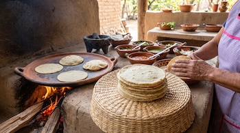 Mujer cocina tortillas de maíz en comal de barro sobre fuego de leña. Hay un molcajete, cuencos con salsas y una pila de tortillas.