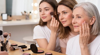Primer plano de tres mujeres de diferentes edades aplicando rubor en sus mejillas, con brochas y compactos de maquillaje sobre un tocador.