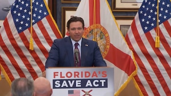 Ron DeSantis, vestido con traje, habla en un atril con un micrófono, flanqueado por las banderas de Estados Unidos y Florida, y un cartel con "FLORIDA'S SAVE ACT"