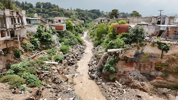 Un río de agua marrón fluye entre dos laderas erosionadas donde hay casas de lámina y concreto construidas cerca del precipicio