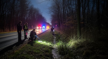 Agentes de policía con linternas inspeccionan una zanja al borde de una carretera rural oscura, con árboles sin hojas y vehículos policiales con luces encendidas al fondo.