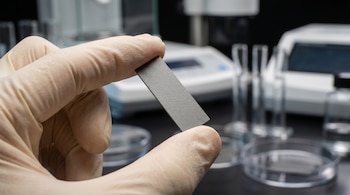 Primer plano de una mano enguantada sosteniendo un pequeño electrodo rectangular gris en un laboratorio con equipo desenfocado.