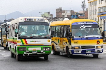 Dos buses de transporte urbano, uno blanco y verde y otro amarillo y azul, circulan en una calle de Lima o Callao, con pasajeros visibles y edificios urbanos de fondo