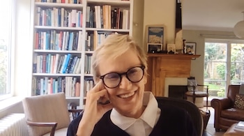 Mujer rubia con gafas y camisa blanca sonríe sentada en una habitación con estantes llenos de libros, un sillón y una chimenea.