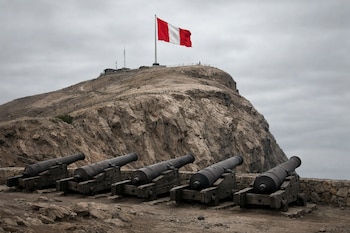 Vista frontal del Morro de Arica con la bandera peruana ondeando en la cima, bajo un cielo nublado, y cinco cañones históricos en primer plano.