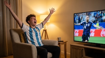 Joven con camiseta de Argentina sentado en sillón, brazos arriba y boca abierta, mira a Lionel Messi en televisión, sala de estar con lámpara.